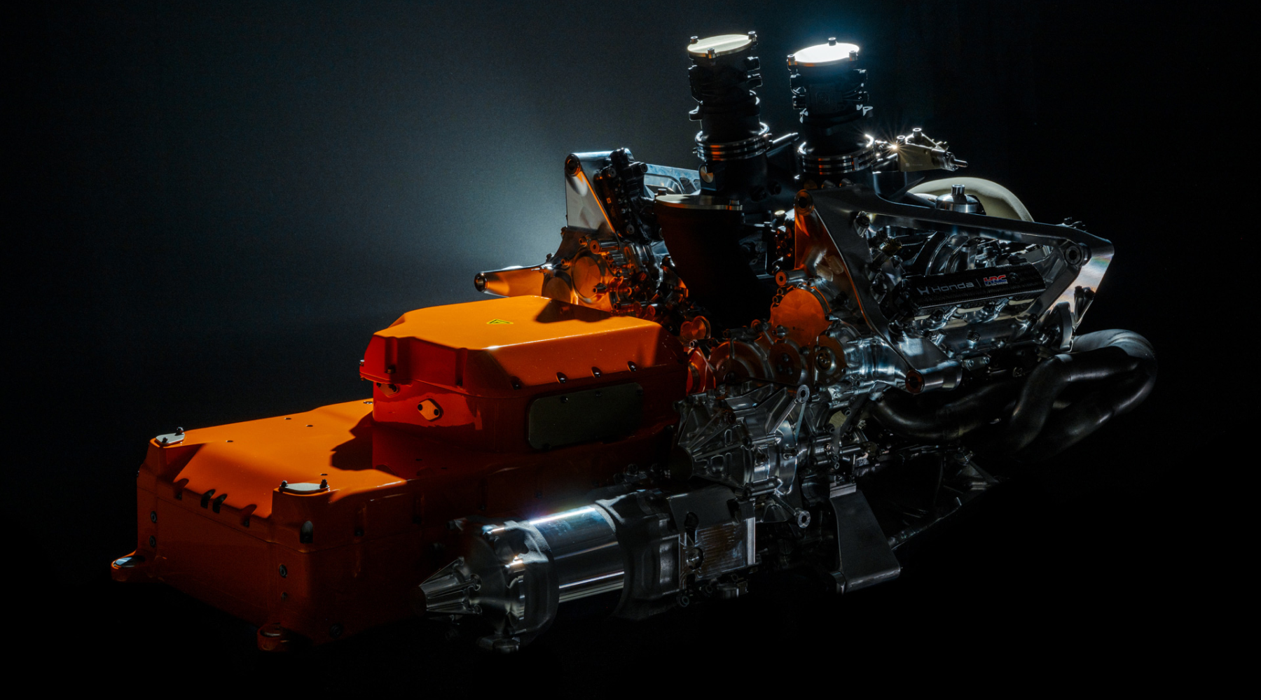 A close-up view of a Honda Formula 1 racing engine, featuring an orange engine block and intricate silver mechanical components. The powerful V6 turbo hybrid power unit is dramatically lit against a dark background, highlighting its complex engineering and design.